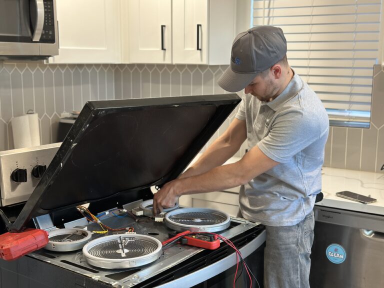 Technician in gray cap repairs an open electric range with burners exposed and wiring visible