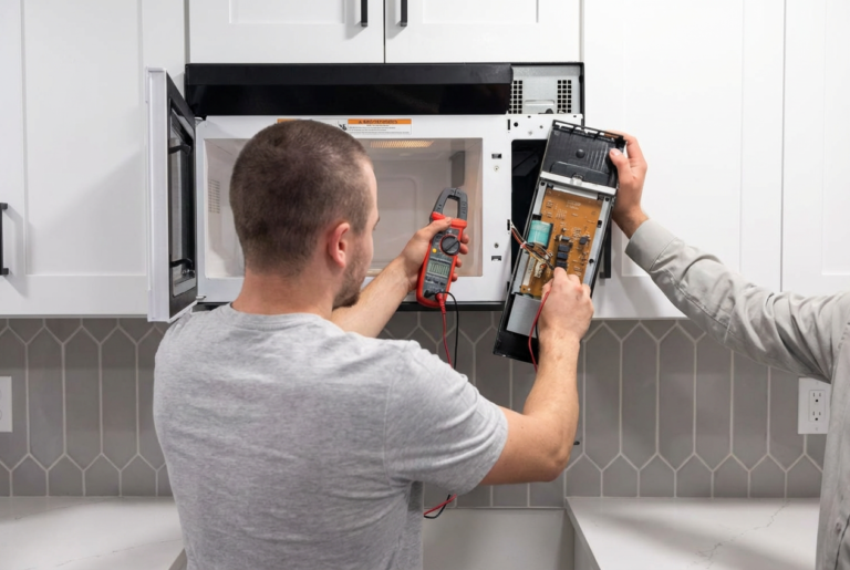 Two technicians repair a microwave; one uses a digital multimeter on the exposed control board while the door is open.